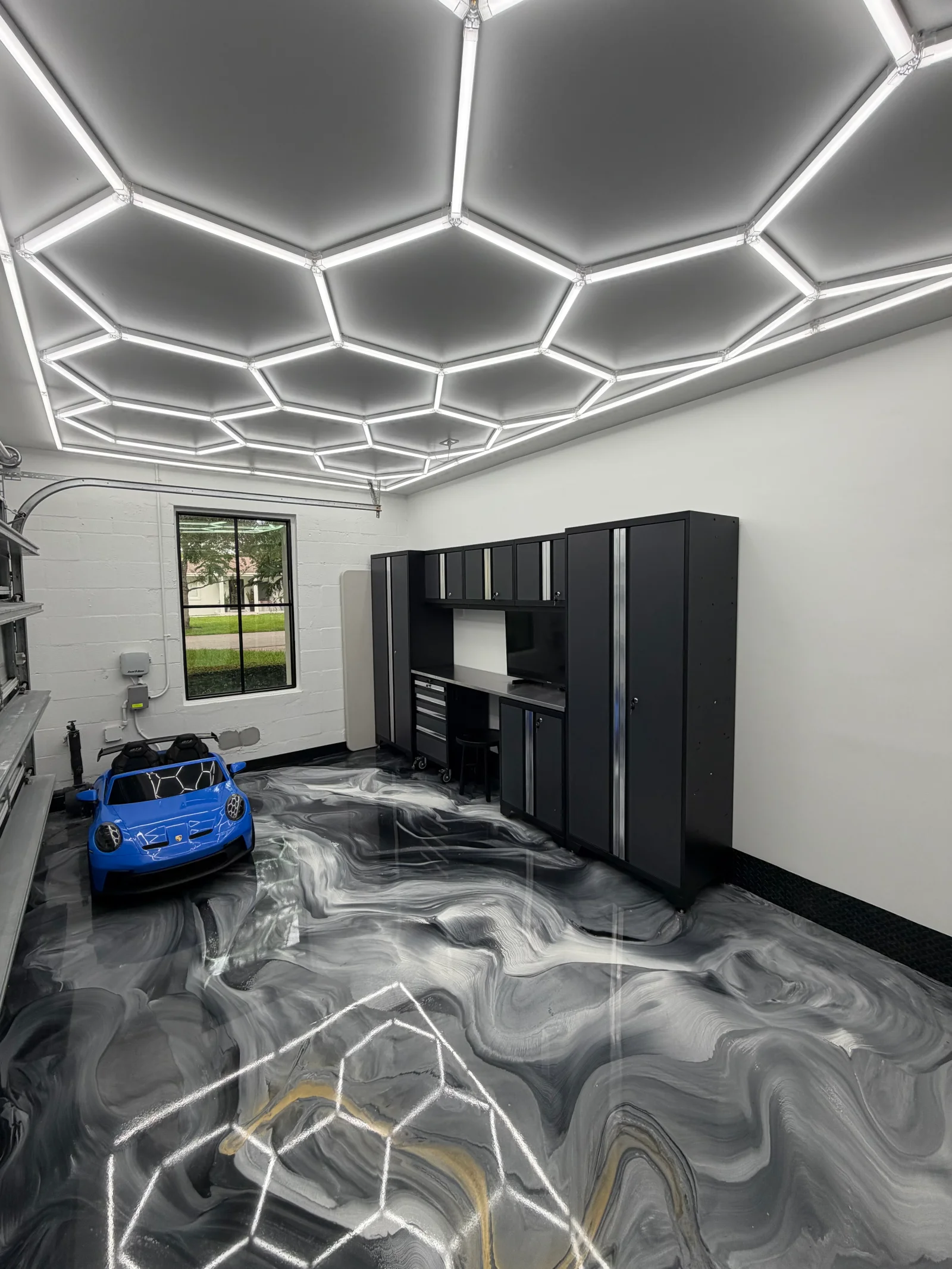 Metallic epoxy garage floor with gray and black swirls, hexagonal LED ceiling panels, and black storage cabinets